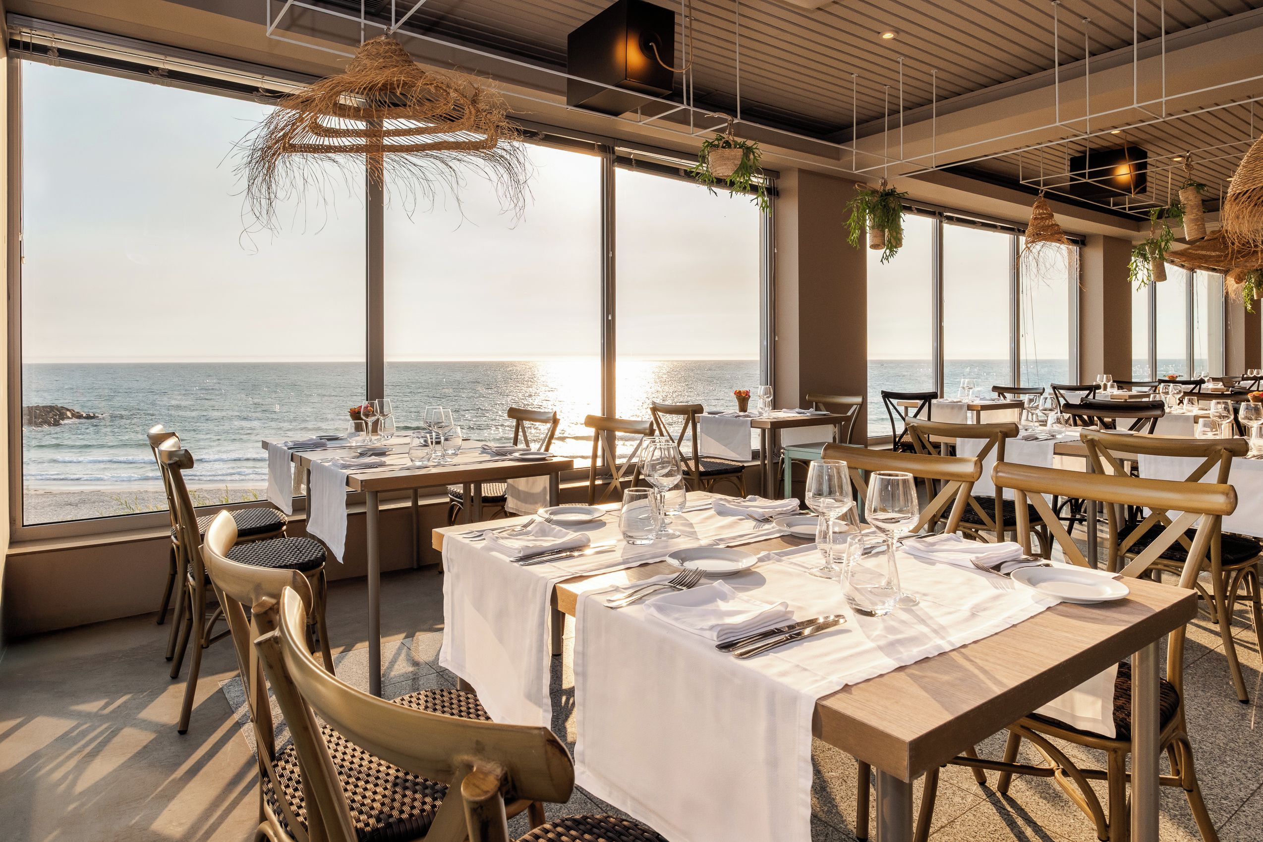 Un restaurante con mesas dispuestas para comer, grandes ventanales con vista al mar y plantas colgantes.