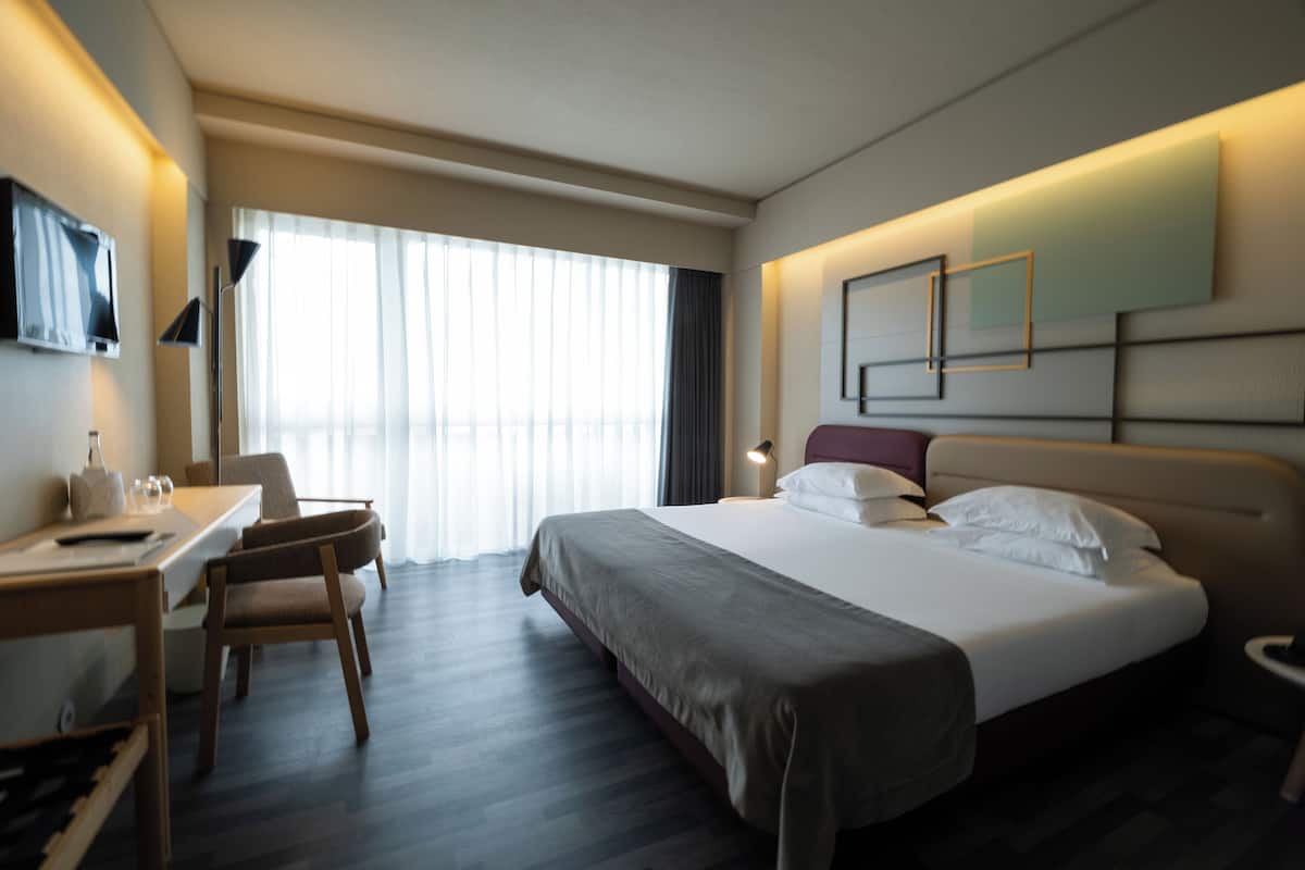 Una habitación de hotel con cama, escritorio, silla y un gran ventanal con cortinas.