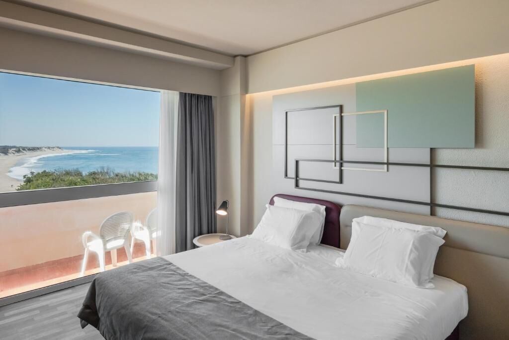A hotel room with a bed, a view of the beach, a chair, and a nightstand.