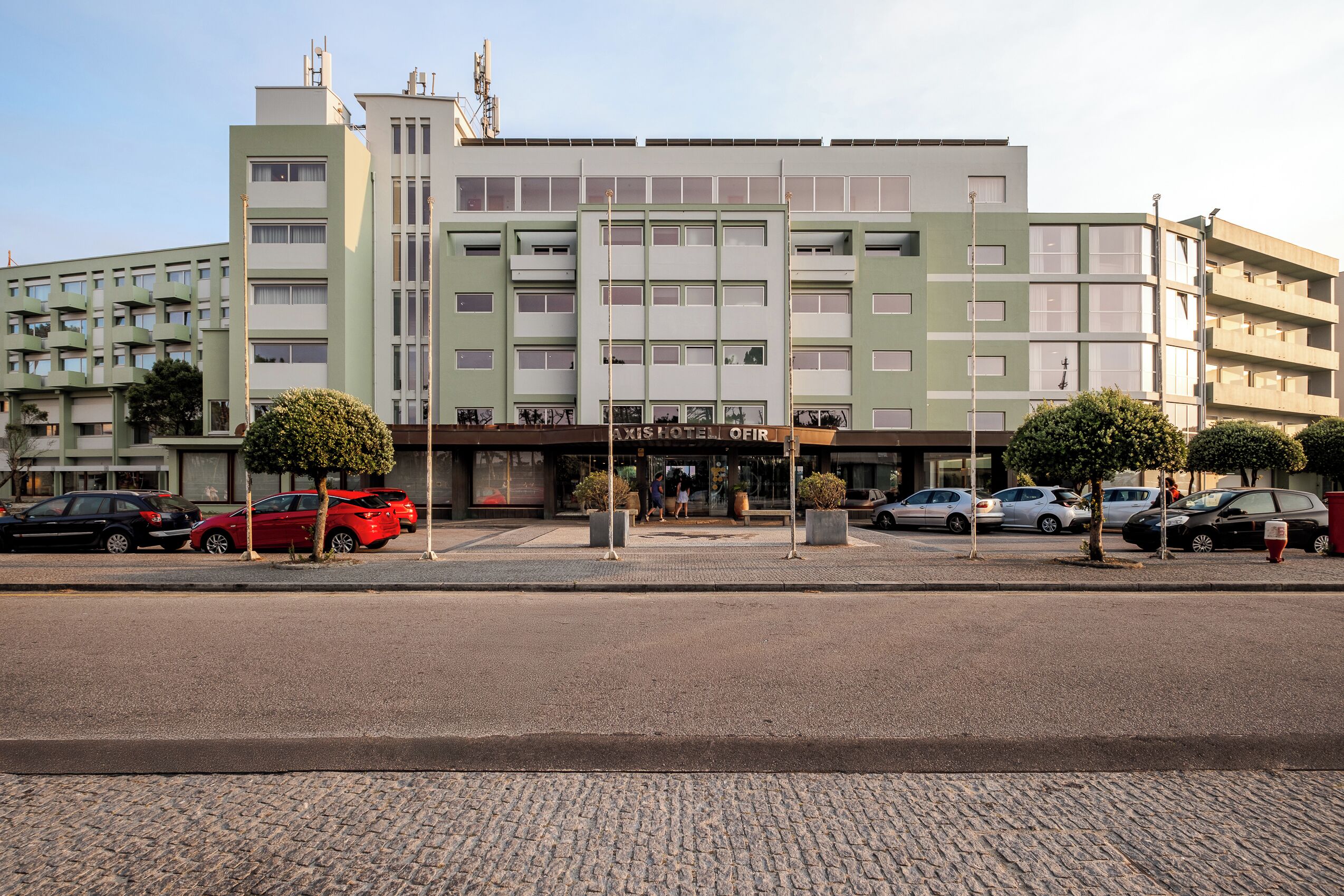 Un hotel de varios pisos con un diseño moderno, que destaca por su entrada principal y varios coches aparcados frente a él.