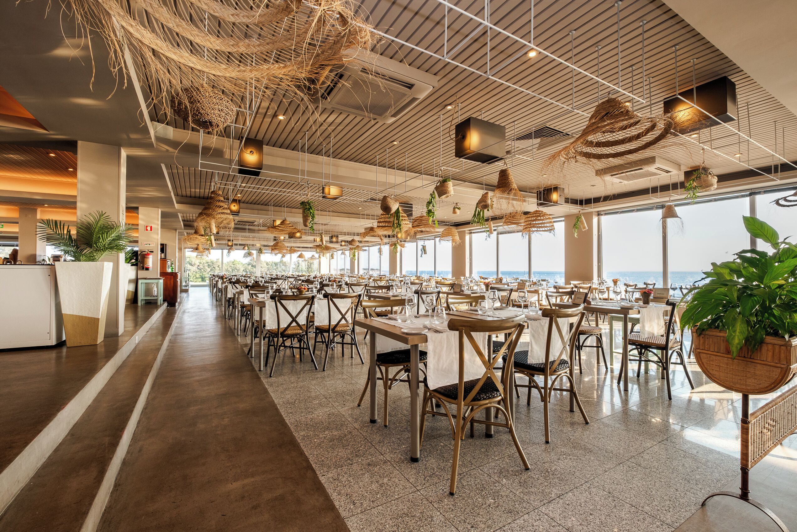 Un comedor espacioso con techos altos, cestas colgantes y grandes ventanales con vistas al mar.