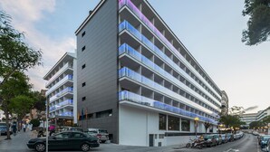 Un hotel de varios pisos con un diseño moderno, que cuenta con balcones en cada planta y un estacionamiento al frente.
