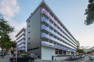 A multi-story hotel with a modern design, featuring balconies on each floor and a parking area in front.