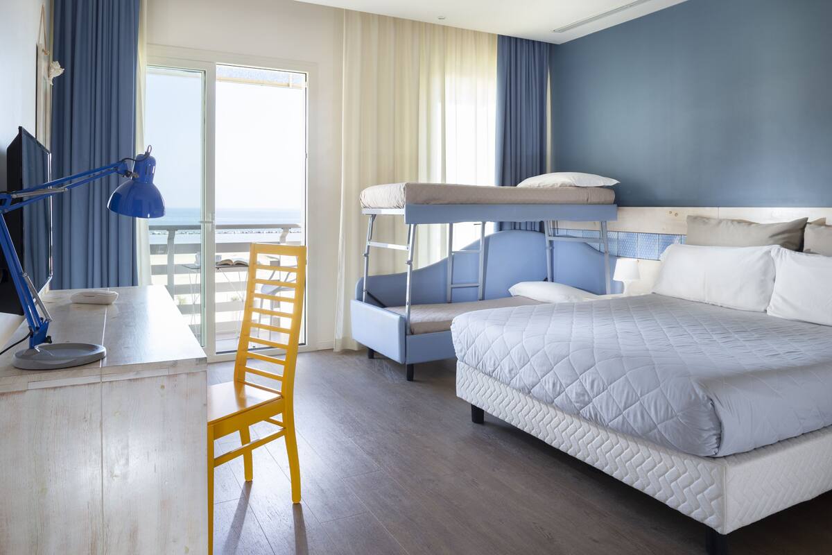 A hotel room with a bunk bed, a desk with a lamp, a chair, and a view of the sea through a large window.