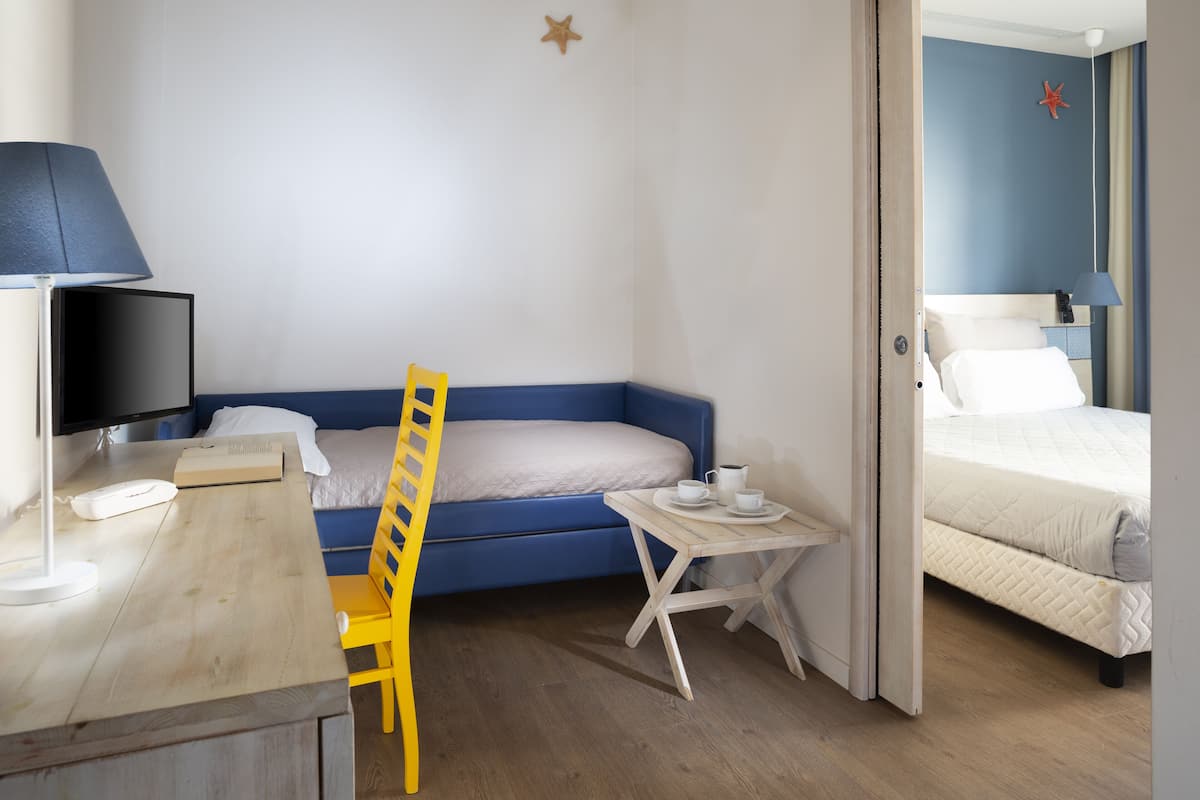 A room with a bed, a desk with a lamp, a chair, and a small table with cups.