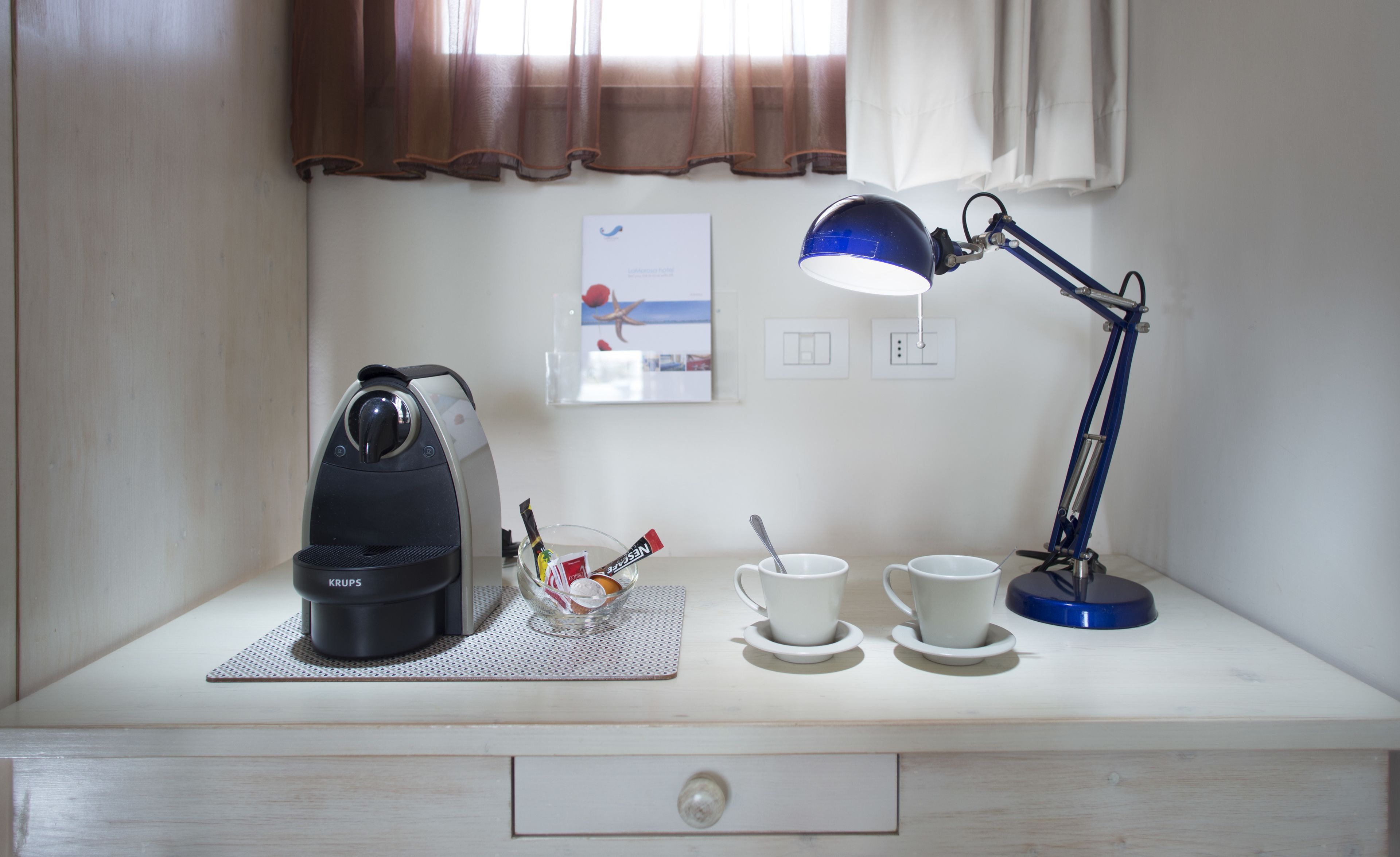 Un escritorio con una cafetera, una taza, un platito, una cuchara, un vaso y una lámpara de escritorio azul.