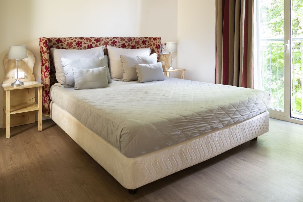 A neatly made bed with a floral headboard, two bedside tables with lamps, and a view of greenery through a window.