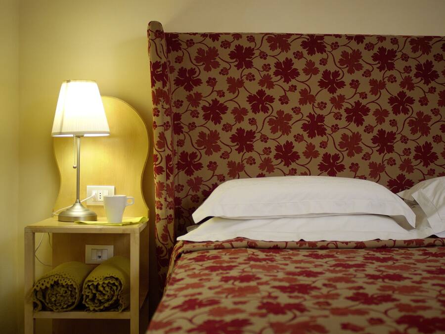 Una cama bien hecha con cabecera estampada con motivos florales, una lámpara de noche y una taza sobre la mesita.