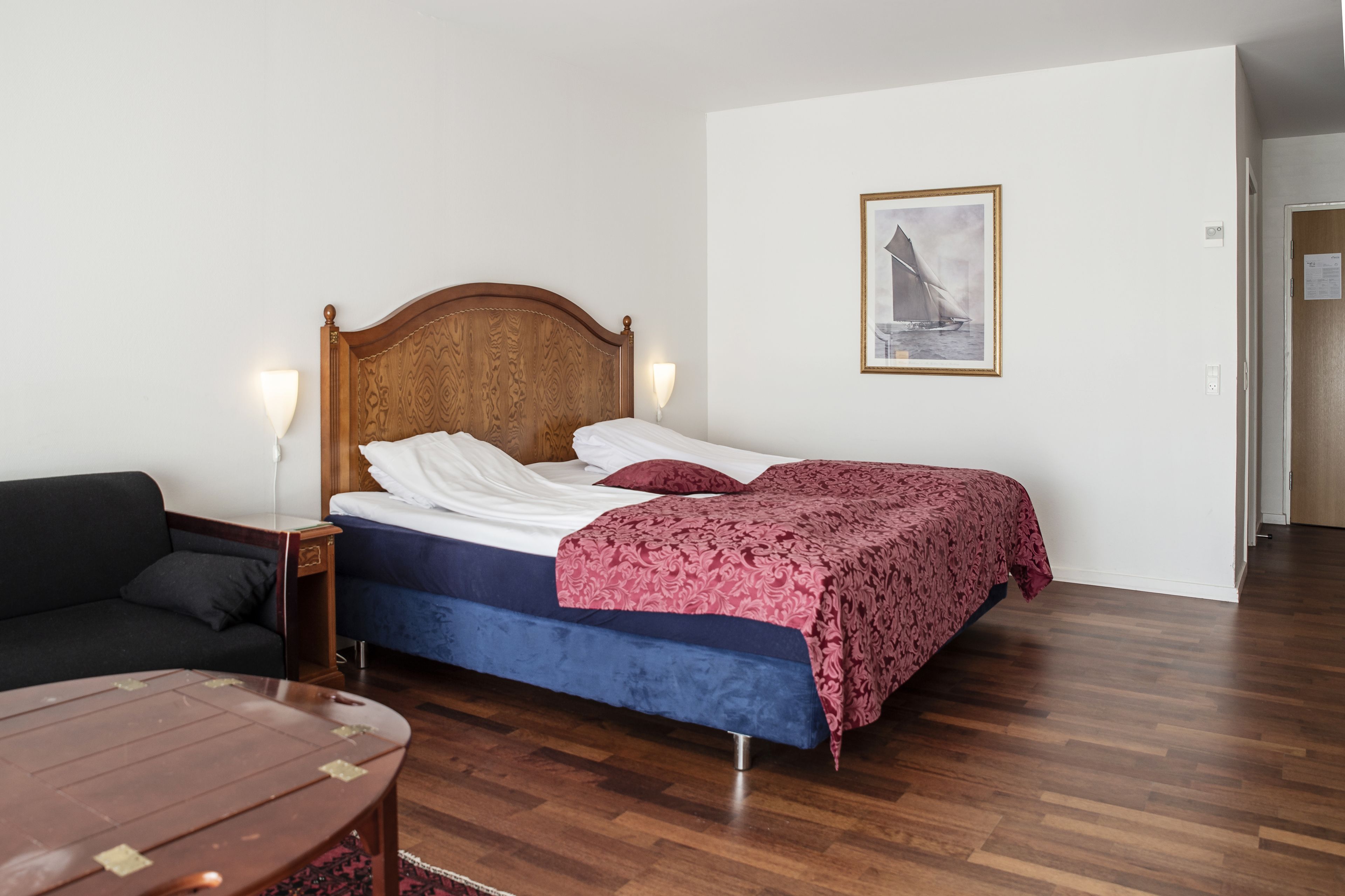A bedroom with a wooden headboard bed, a blue mattress, a red and white patterned blanket, a black sofa, and a wooden table.