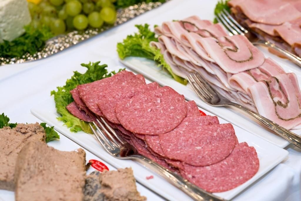 A buffet table with various meats, including sliced ham and salami, arranged on white plates with lettuce and grapes.