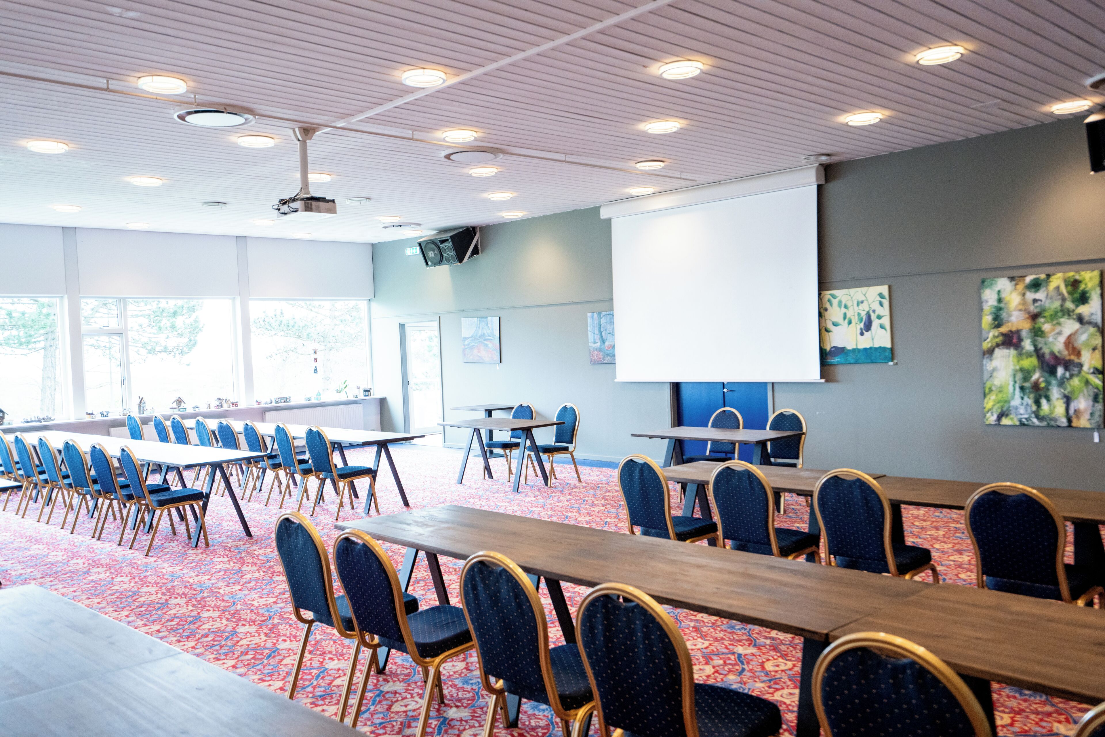 Ein Konferenzraum mit langen Tischen und Stühlen, einer Projektionsleinwand und Kunstwerken an den Wänden.