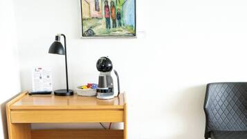 A wooden desk with a black lamp, a coffee maker, and a basket. A black chair is to the right.