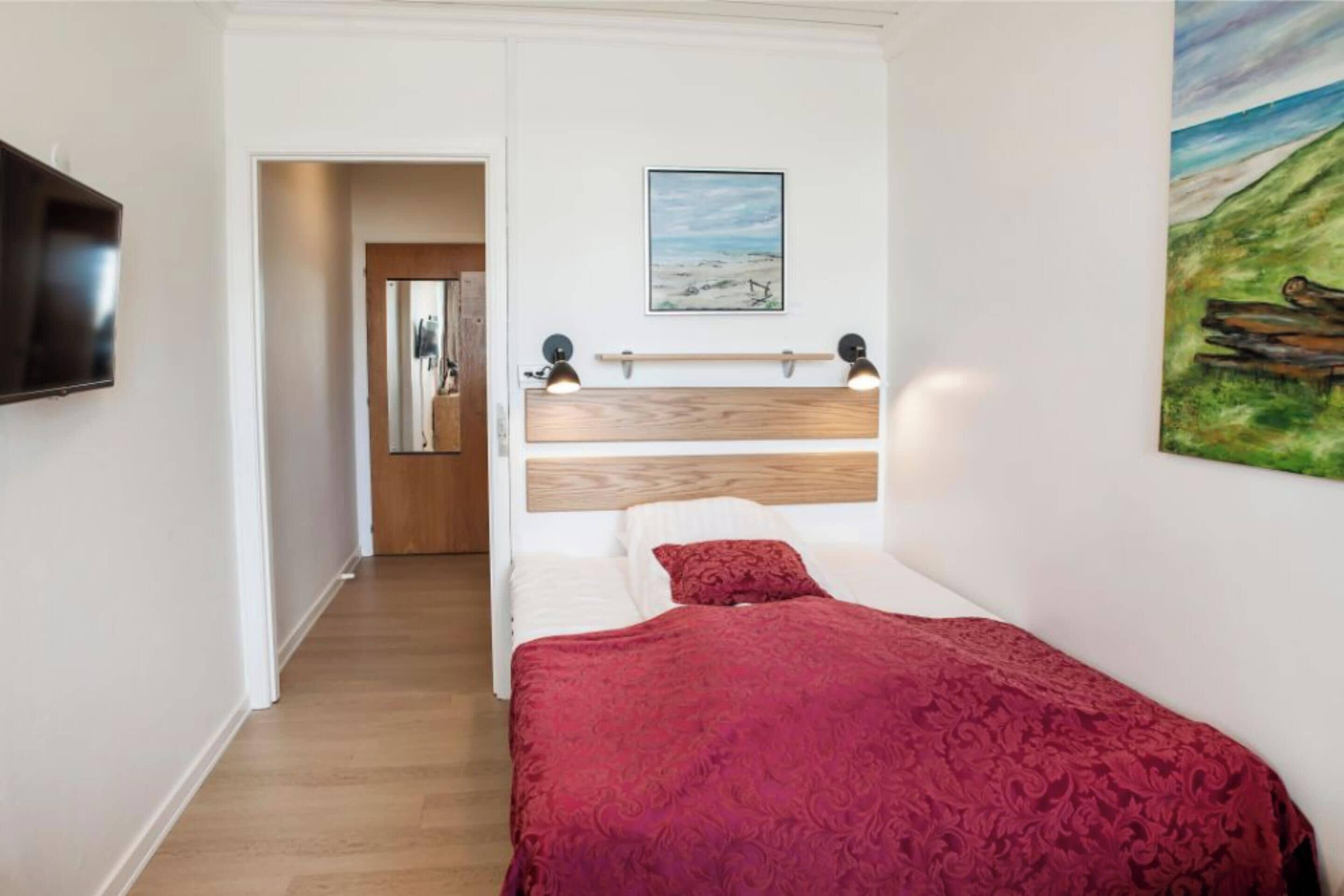 A bedroom with a wooden headboard, a red bedspread, a painting of a landscape, and a mounted television.