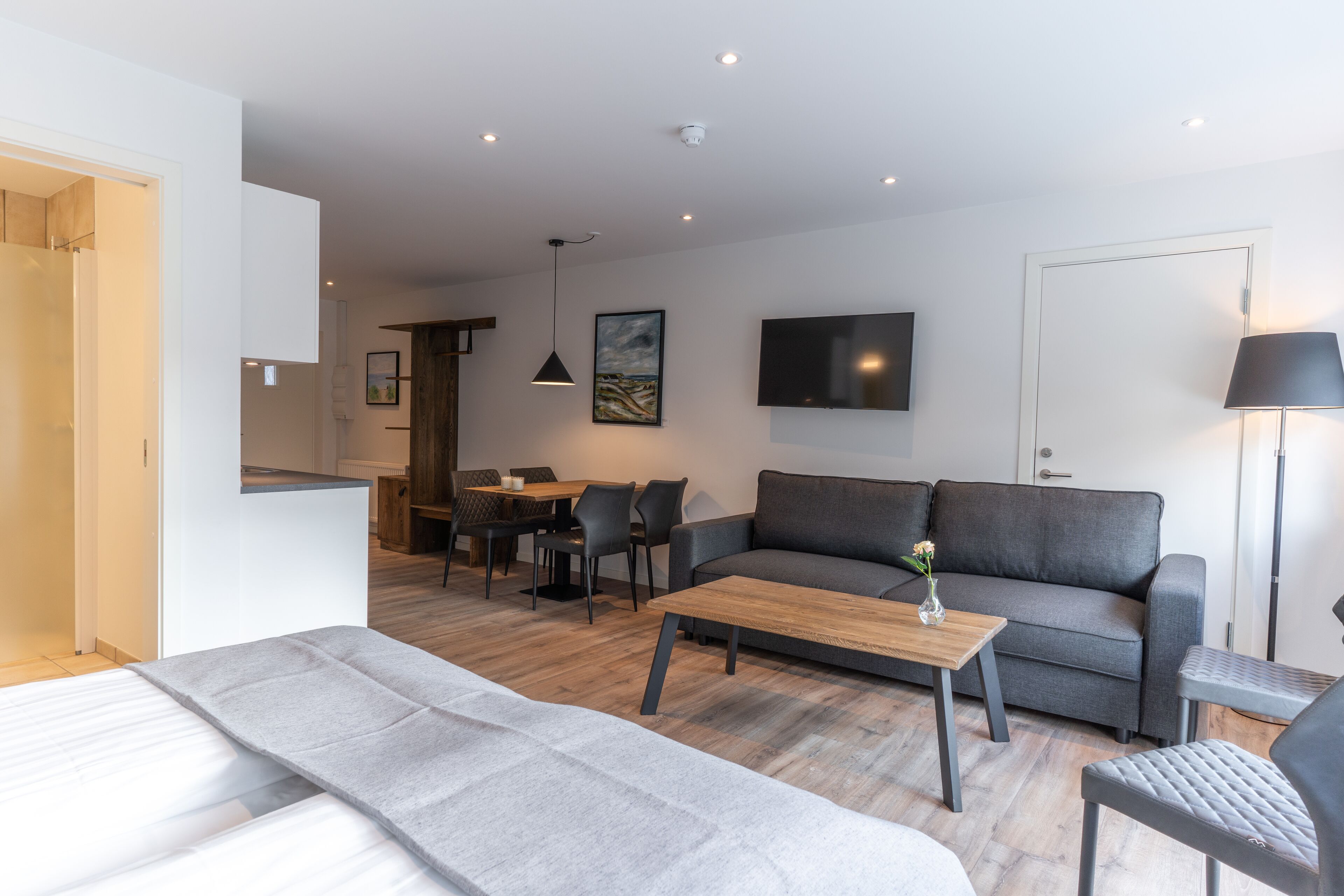A modern living room with a grey sofa, a wooden coffee table, and a grey armchair. There is a dining area with a table and chairs in the background.