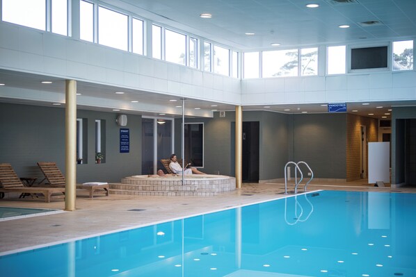 An indoor swimming pool with a sunlit atrium, lounge chairs, and a spa area. - Hotel Marina (Grenaa)
