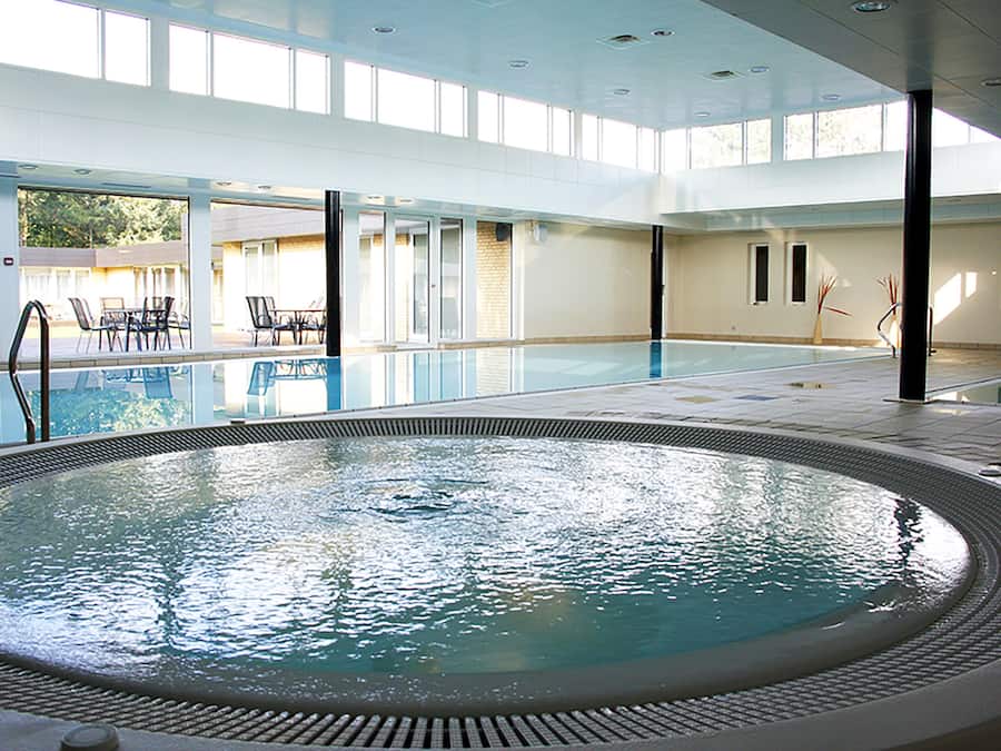 An indoor swimming pool with clear water, surrounded by lounge chairs and tables, and large windows allowing natural light.