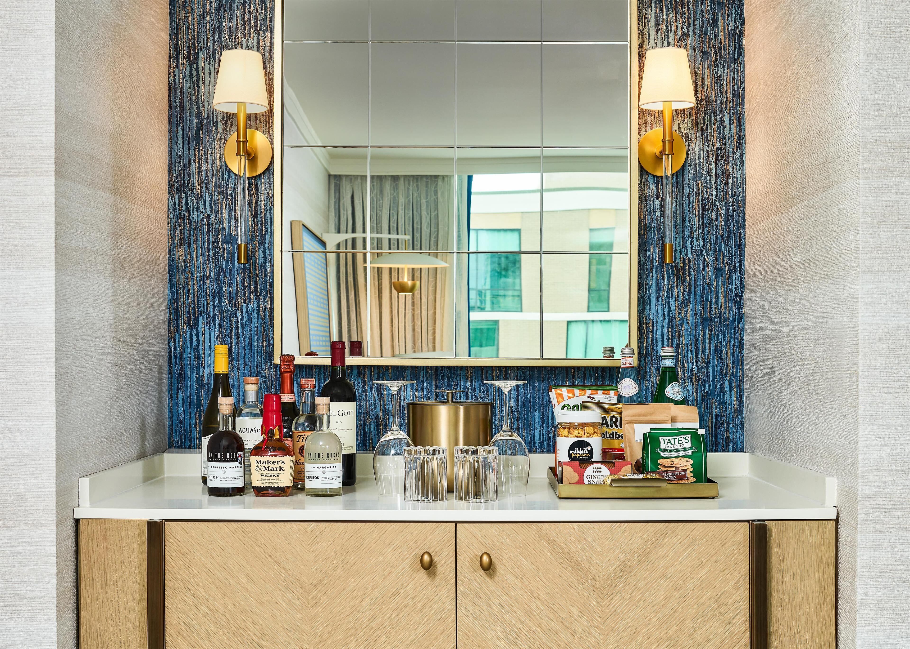 A bathroom vanity with a mirror, two wall-mounted lamps, and a variety of toiletries and beverages.