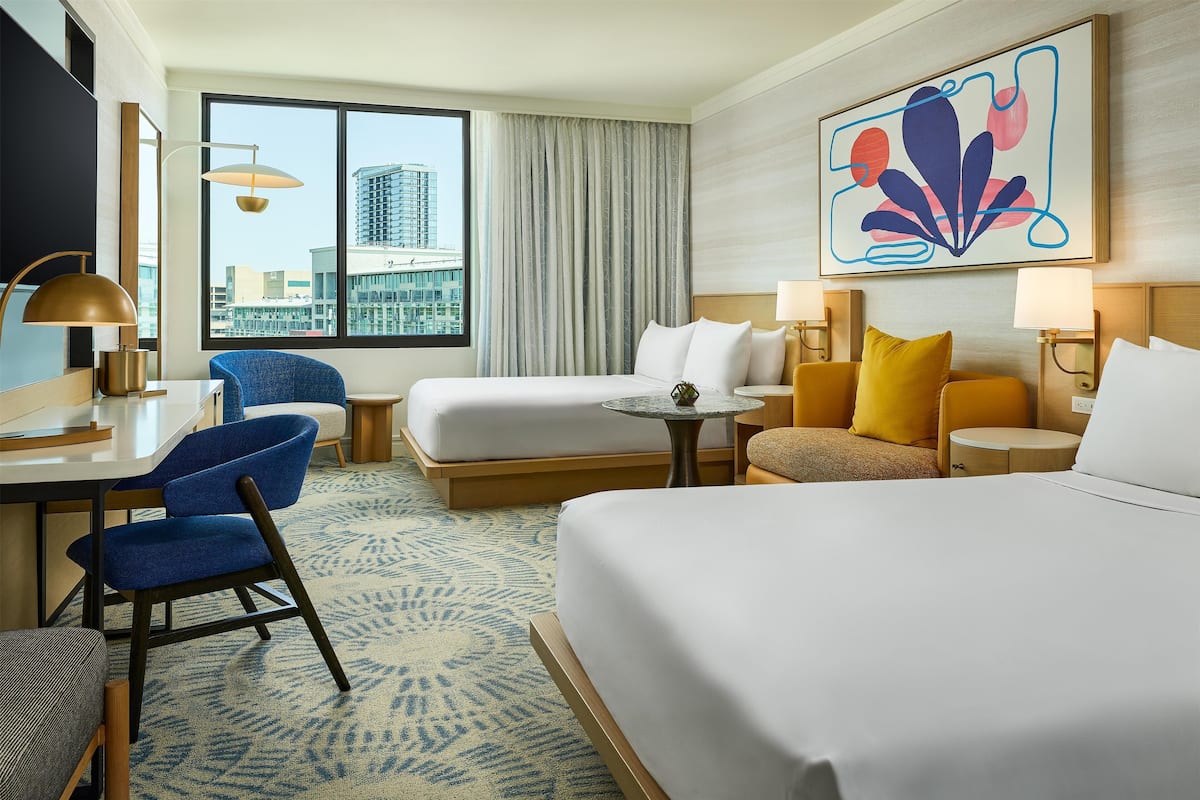 A hotel room with a bed, a desk with a chair, a small table, and a sofa. There is a large window with a view of buildings.