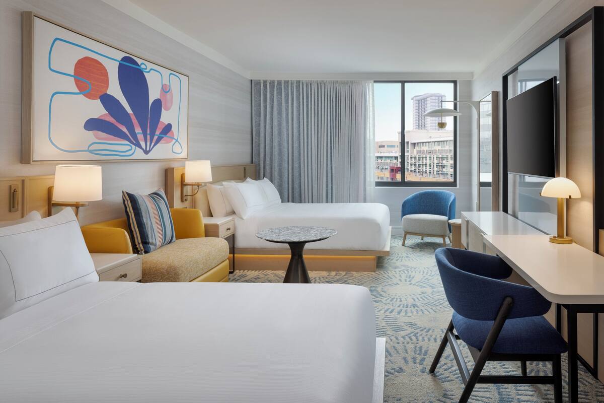 A hotel room with a bed, a desk, a chair, a small table, and a large window with a city view.