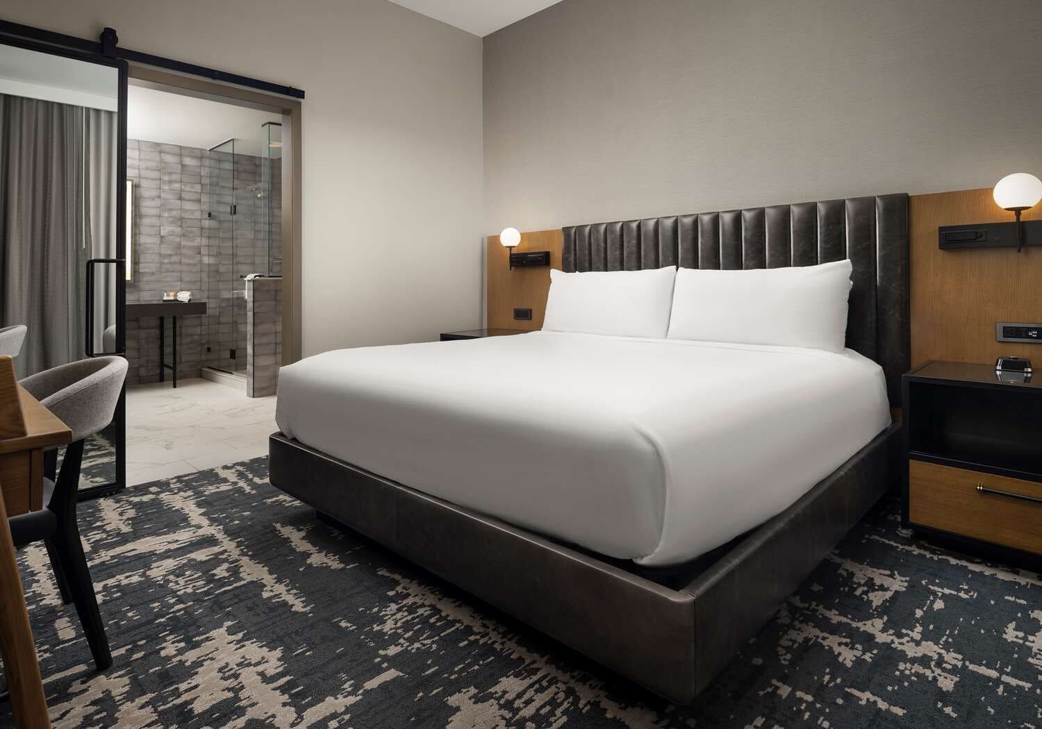 A modern hotel room with a large bed, a desk, a chair, a shower area, and a bathroom.