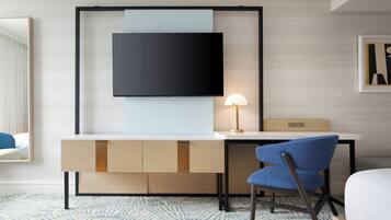 A modern hotel room with a flat-screen TV mounted on a wall, a desk with a lamp, a blue chair, and a bed.