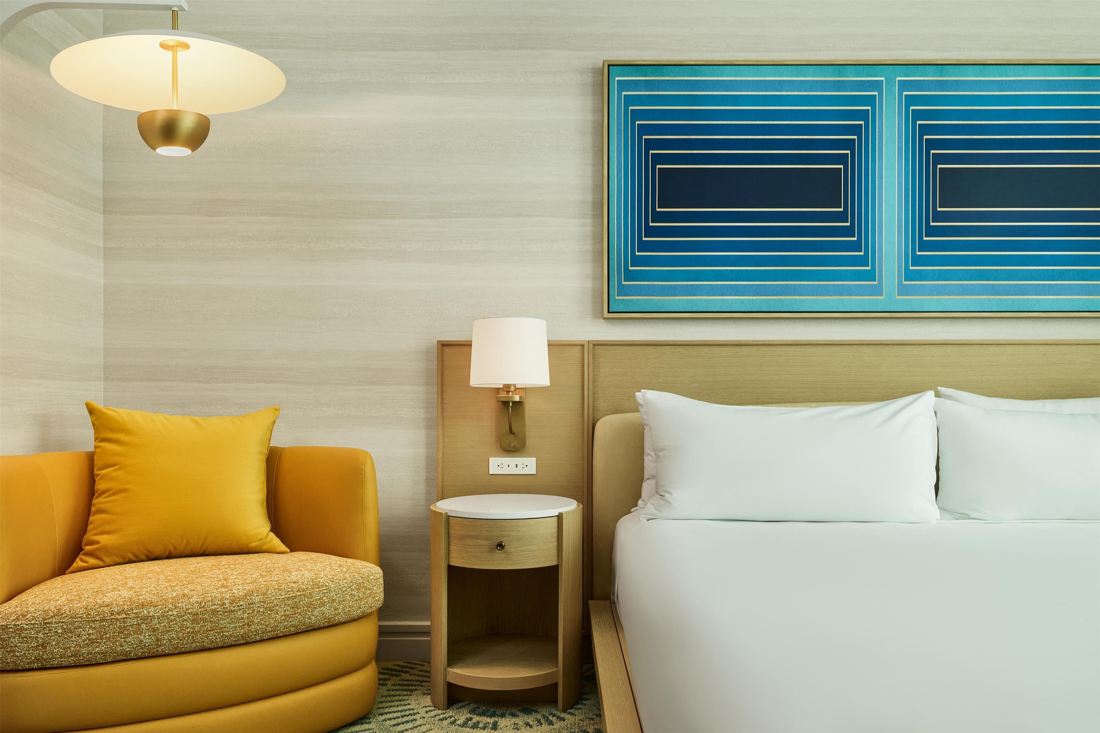 Habitación de hotel con cama, mesita de noche con lámpara, sofá con almohada y una obra de arte en la pared.