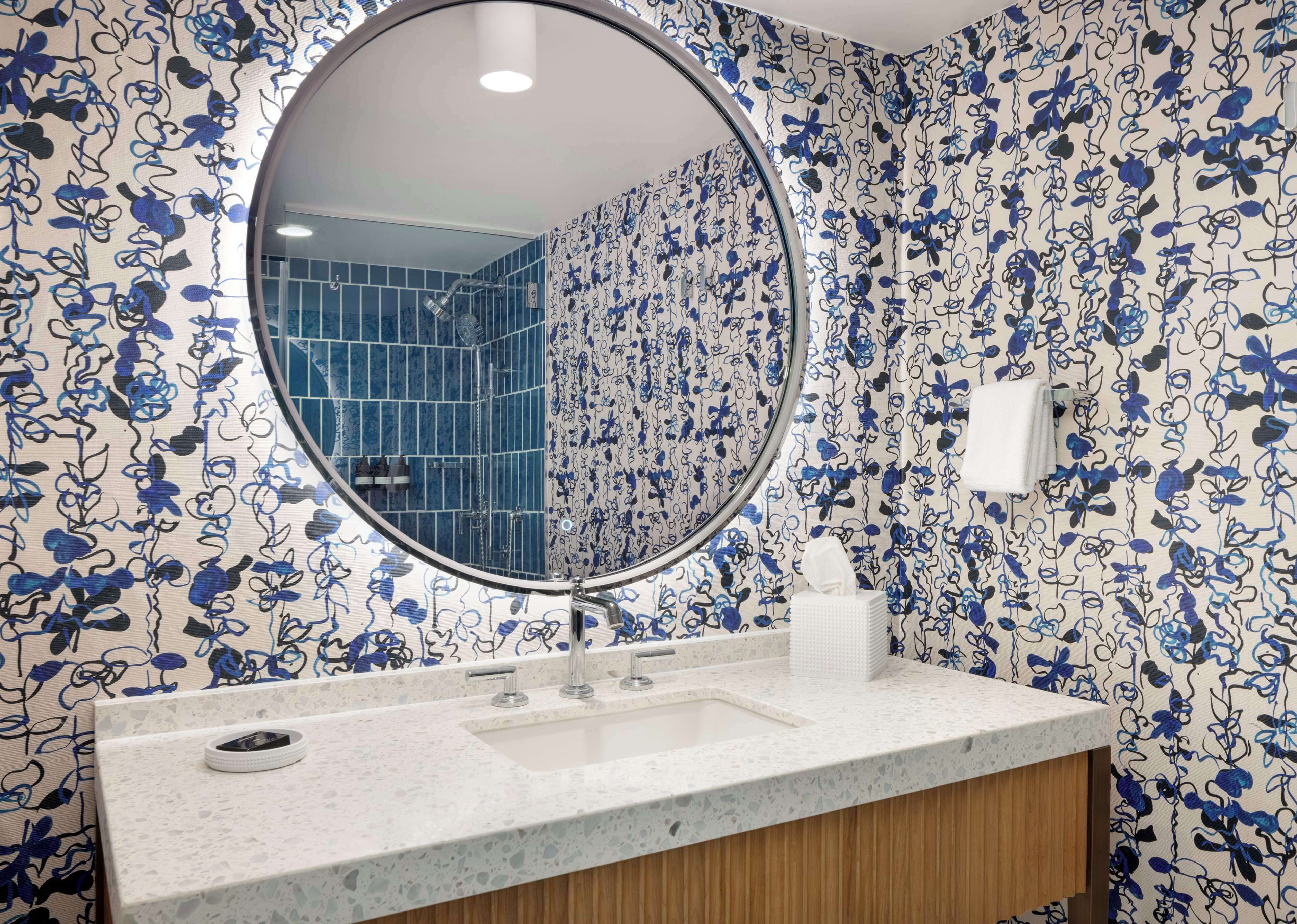 Un baño con espejo redondo, lavamanos blanco y papel tapiz con motivos florales azules.