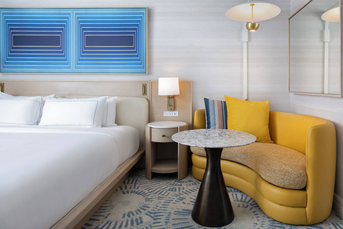 A hotel room with a bed, a yellow sofa, a small round table, a nightstand, a lamp, and two blue artwork pieces on the wall.