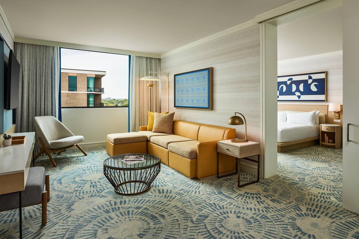 A modern hotel room with a sectional sofa, a round coffee table, a bed, and a view of a building outside.