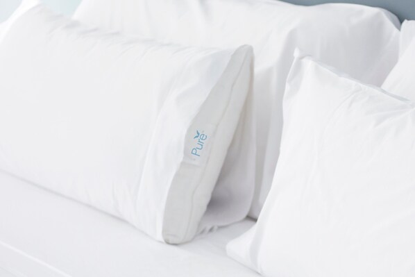 Una cama con ropa de cama y almohadas blancas, una de ellas con un nombre de marca visible.