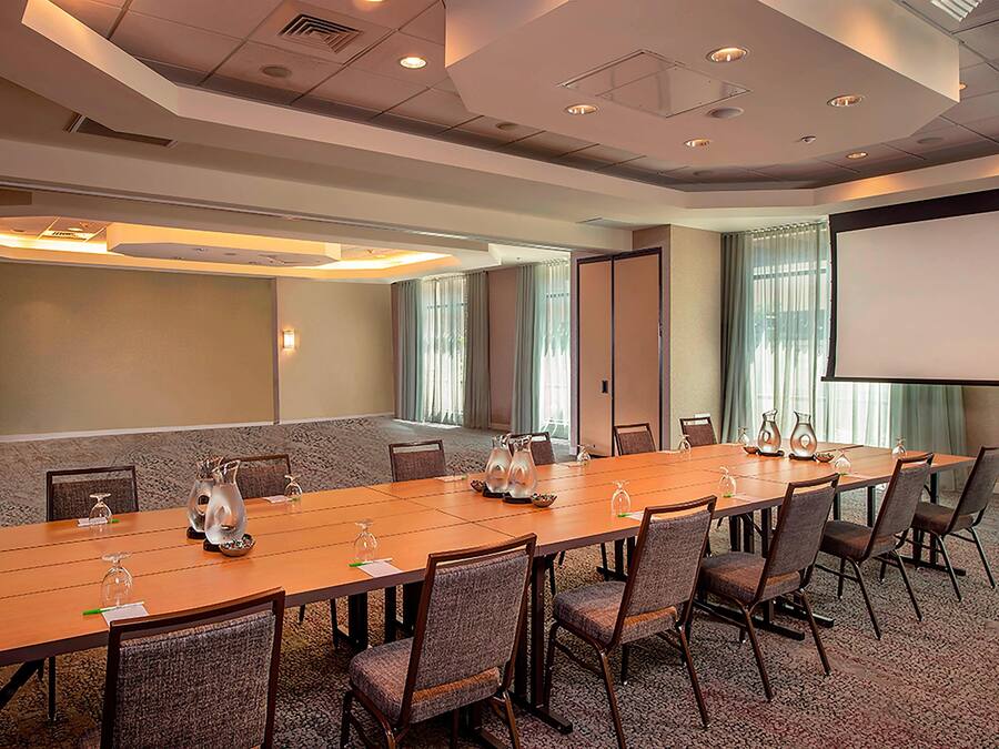 A conference room with a long table, chairs, a projector screen, and a large window with curtains.