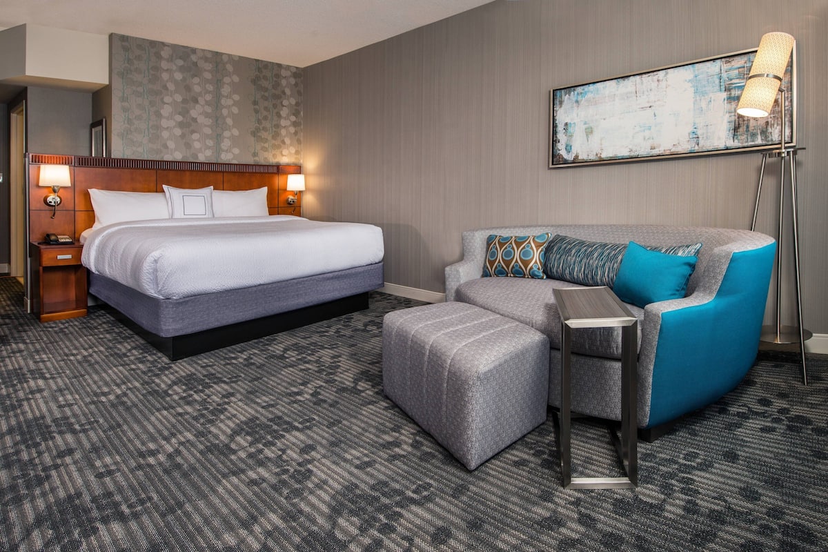 Habitación de hotel con una cama grande, un sofá gris, un pequeño otoman, una lámpara de pie y una pared con diseño texturizado.