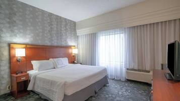 Habitación de hotel con una cama grande, una mesita de noche con un reloj, un televisor y una ventana con cortinas.