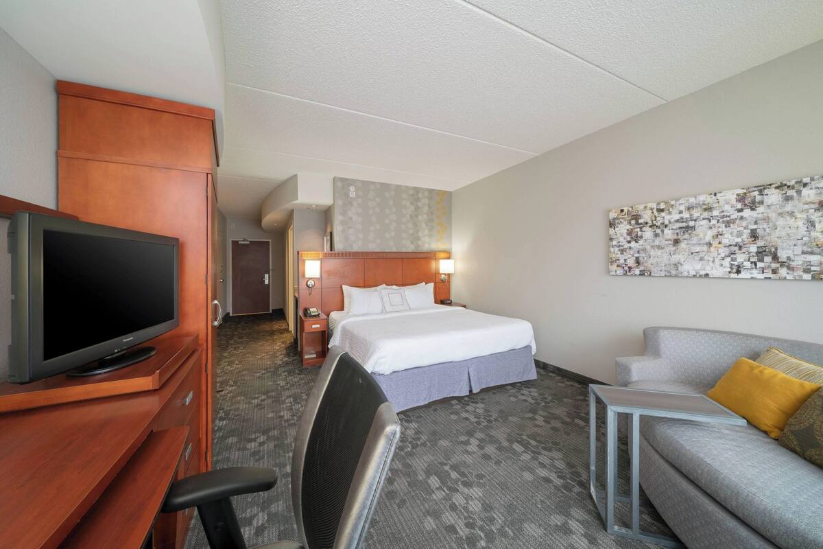 A hotel room with a bed, a TV, a desk, a chair, a sofa, and a painting on the wall.