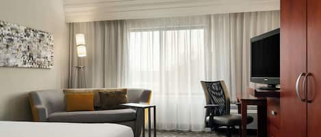 A hotel room with a bed, a sofa, a desk with a television, a chair, and a large window with curtains.