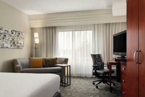 Habitación de hotel con cama, sofá, escritorio con televisor, silla y un amplio ventanal con cortinas.