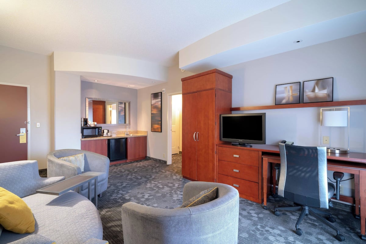 A hotel room with a sofa, armchair, desk, TV, and kitchenette.