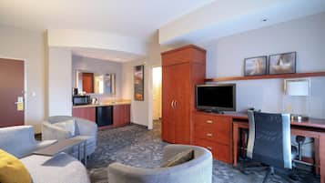 A hotel room with a sofa, armchair, desk, TV, and kitchenette.