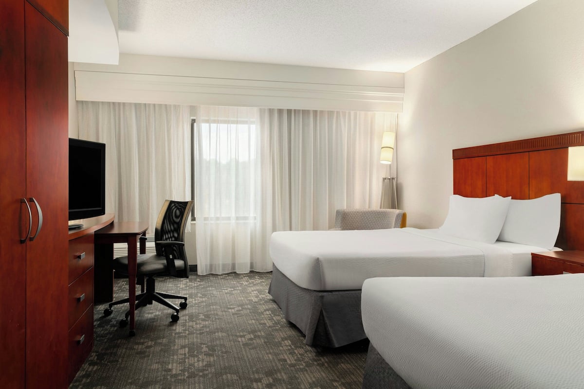 A hotel room with two beds, a desk with a television, a chair, and a lamp.