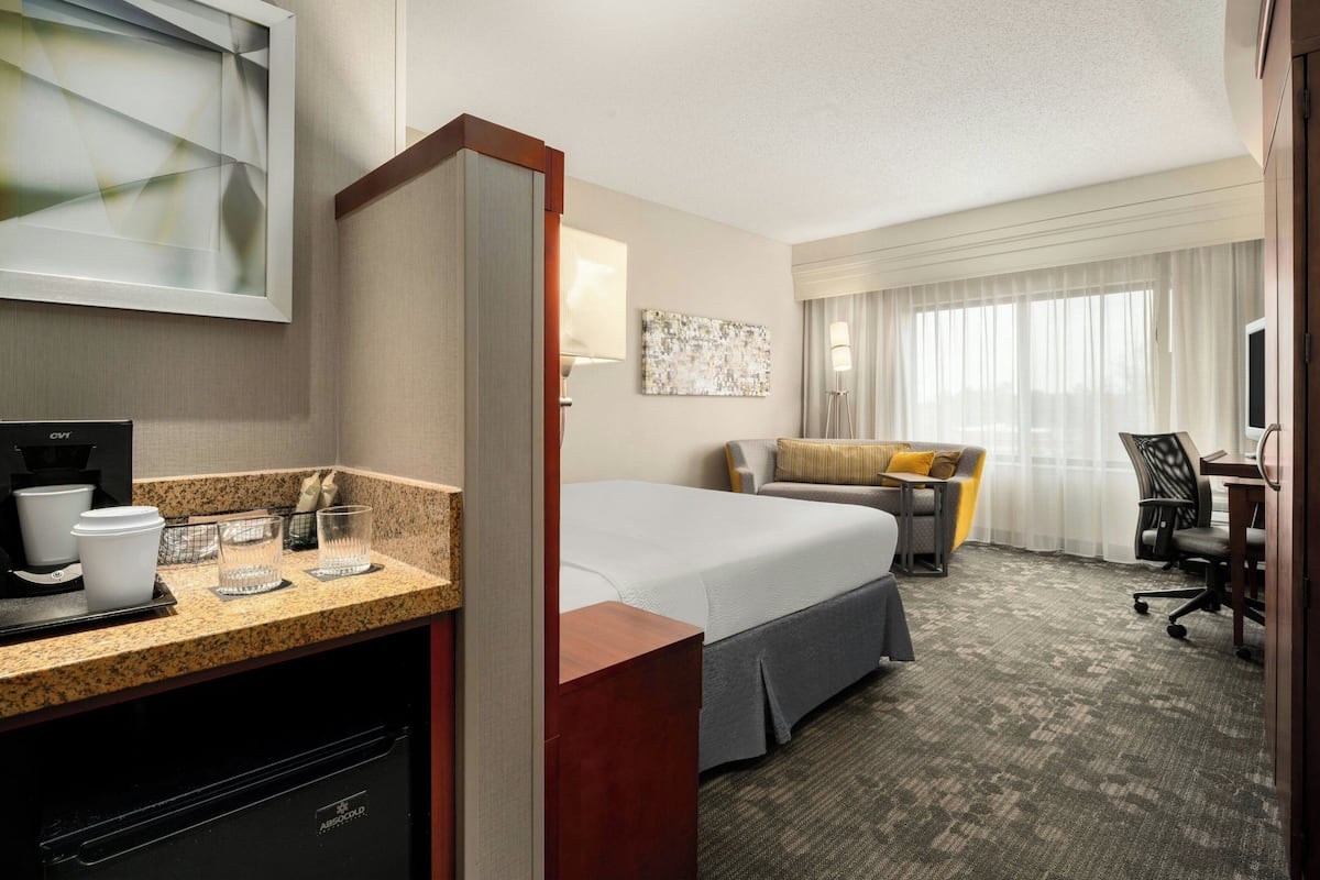 A hotel room with a bed, a desk, a chair, a coffee machine, and a window with curtains.
