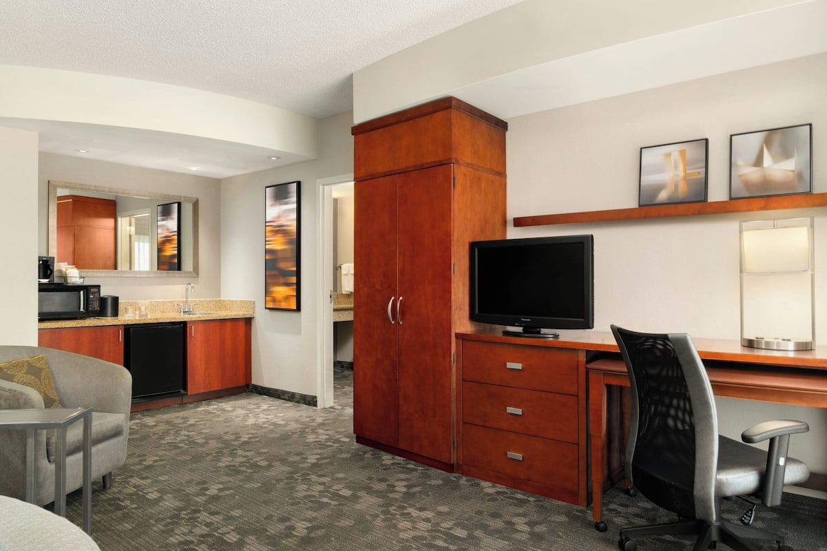 A hotel room with a kitchenette, a desk with a computer, a TV, a chair, and a large wardrobe.