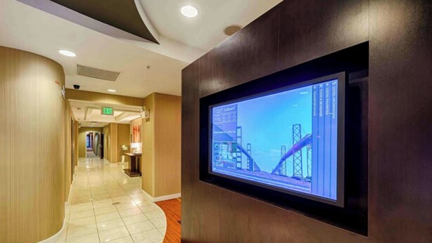 Pasillo de hotel con una pantalla grande mostrando una obra en construcción, un muro curvo de madera y un letrero de salida.