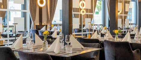 Un restaurante moderno con sillas de terciopelo oscuro, mesas ordenadas con servilletas blancas y ventanas amplias con cortinas.