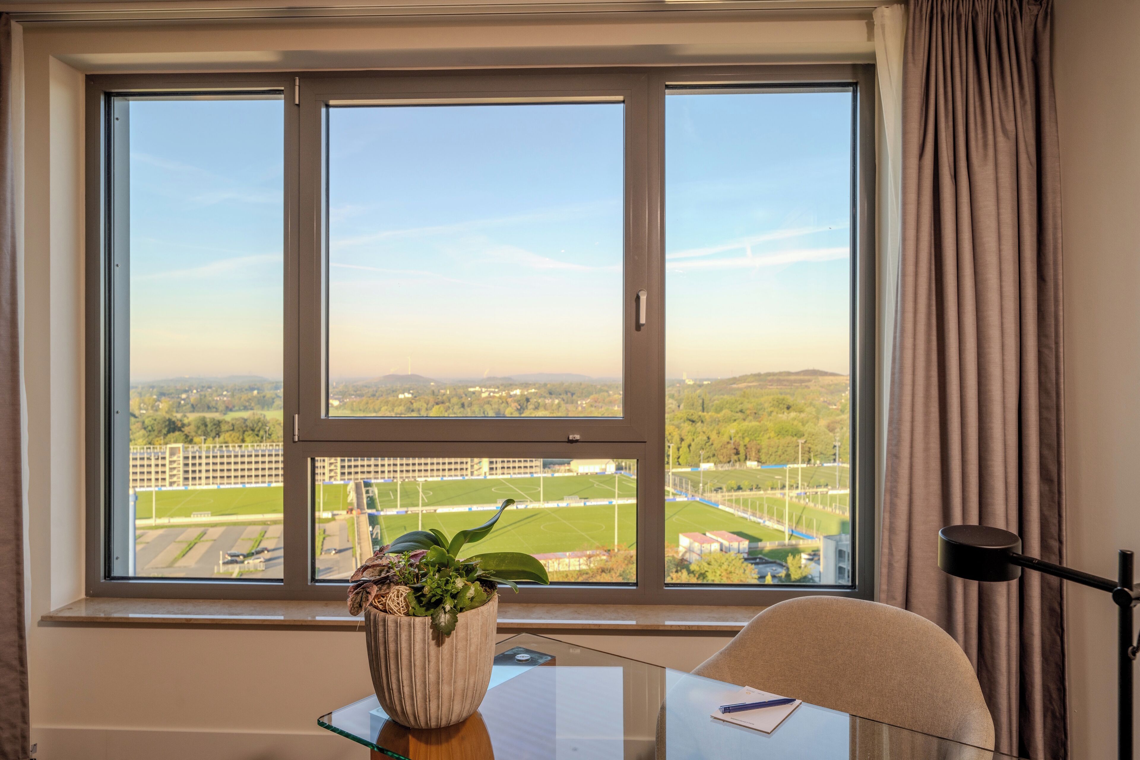 Ein Zimmer mit großem Fenster und Blick auf ein Sportfeld sowie die Ferne.