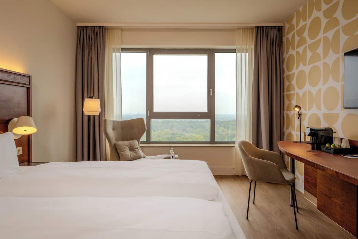 A hotel room with a large bed, two chairs, a desk, and a large window offering a view.