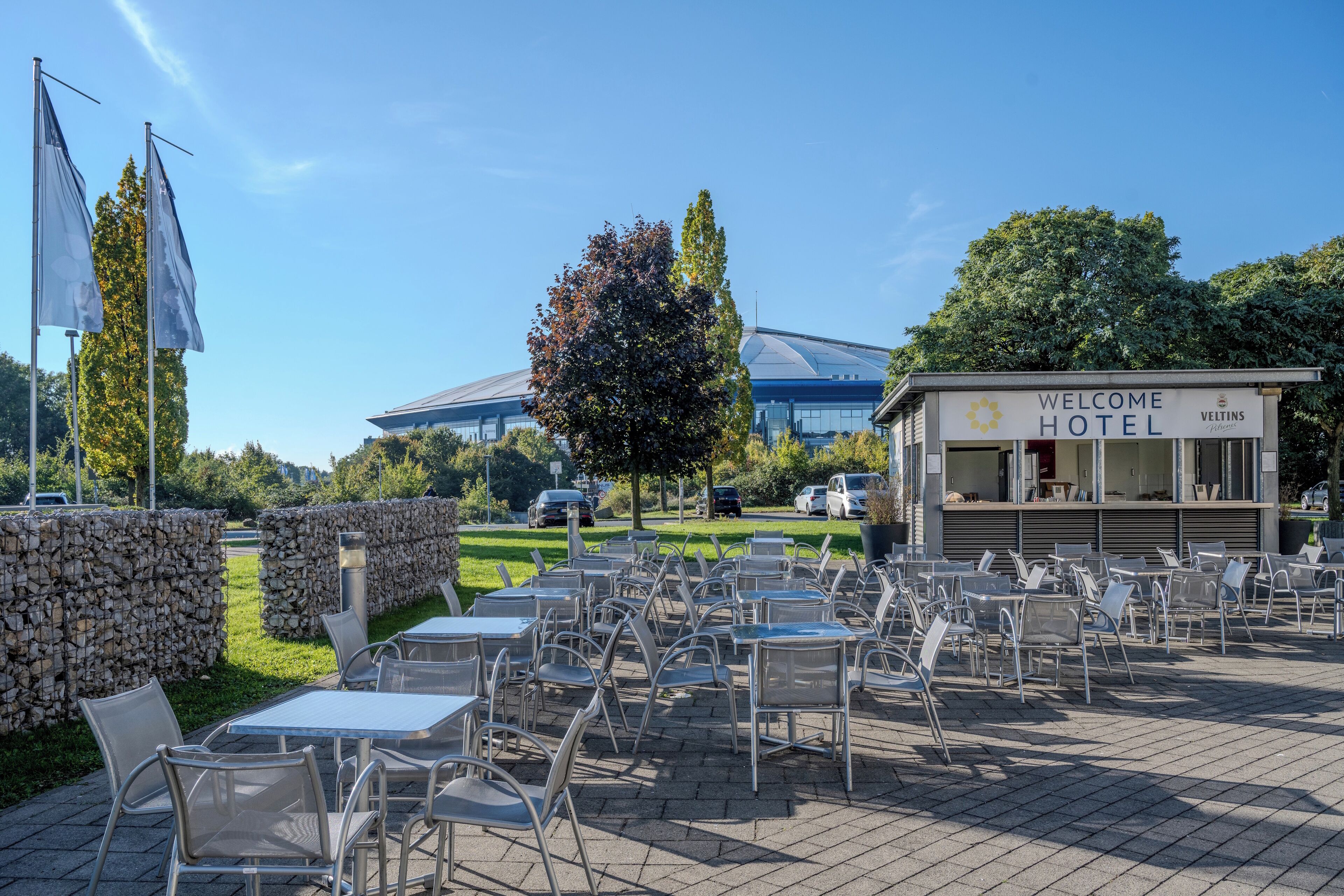An outdoor seating area with tables and chairs, a sign for the Welcome Hotel, and a modern building in the background.
