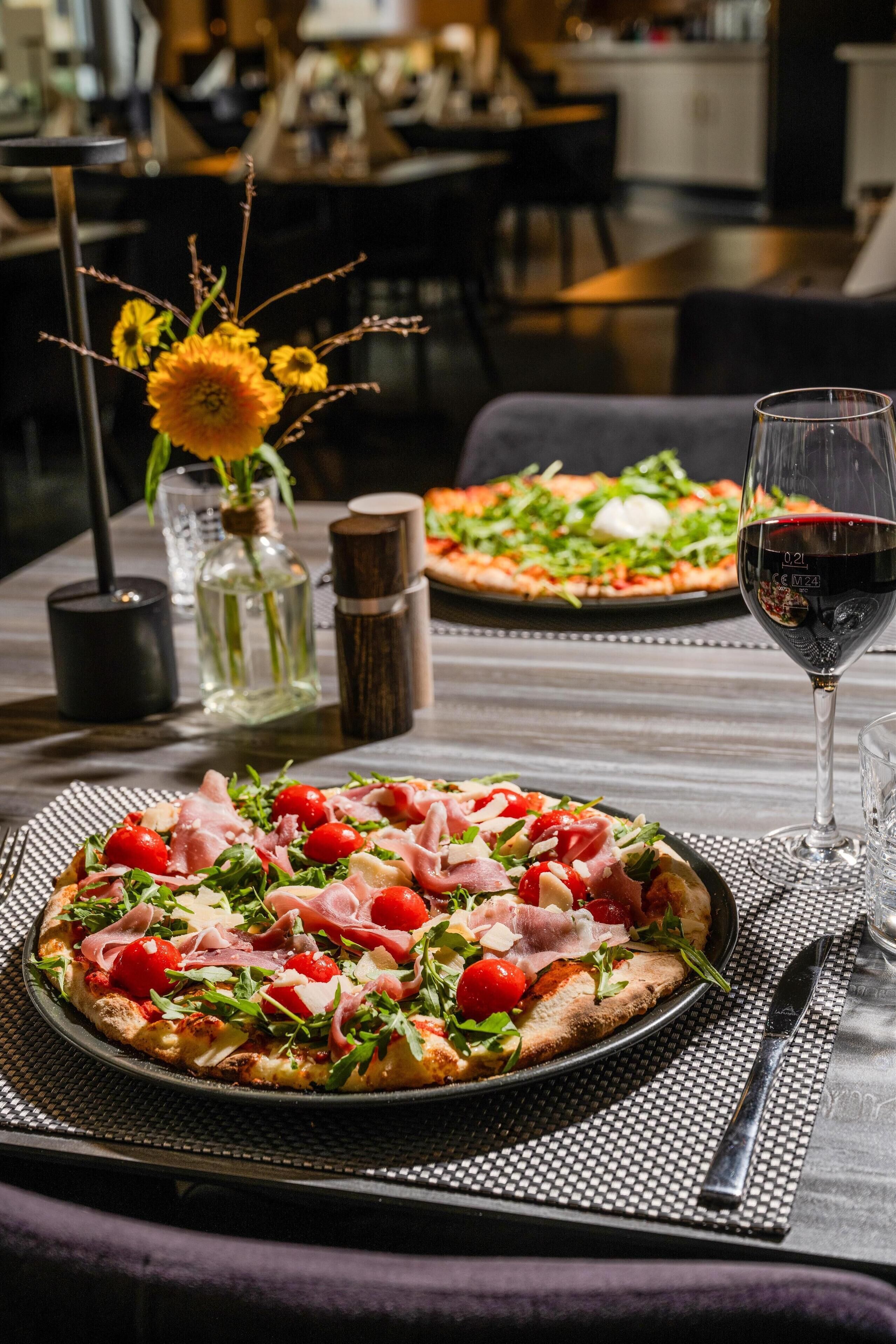 Una pizza con diferentes ingredientes, una copa de vino tinto y un jarrón con flores sobre una mesa.