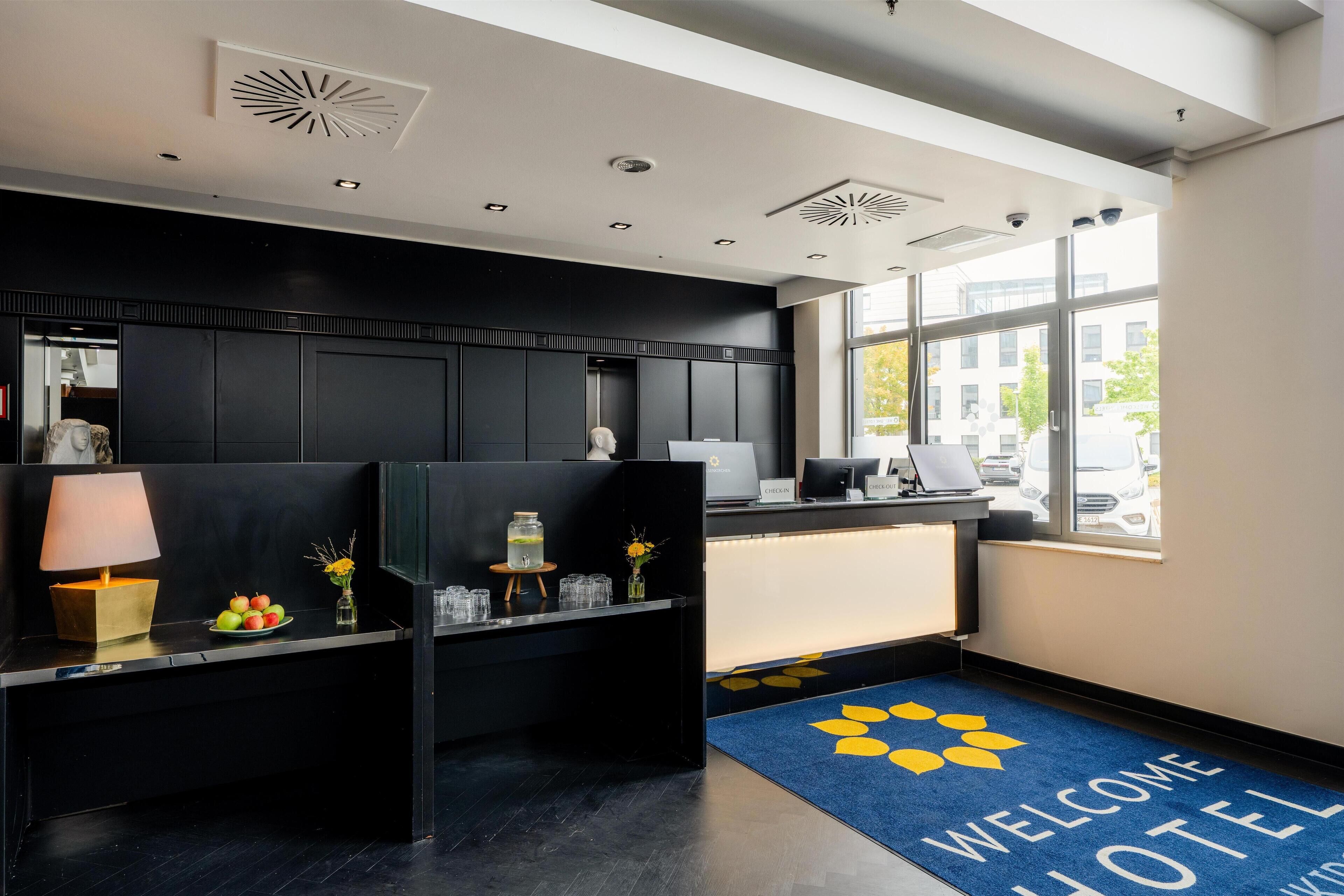 Ein Hotel-Rezeptionsbereich mit blauem Teppich, einem Empfangstresen, einer Lampe und einer Obstschale.