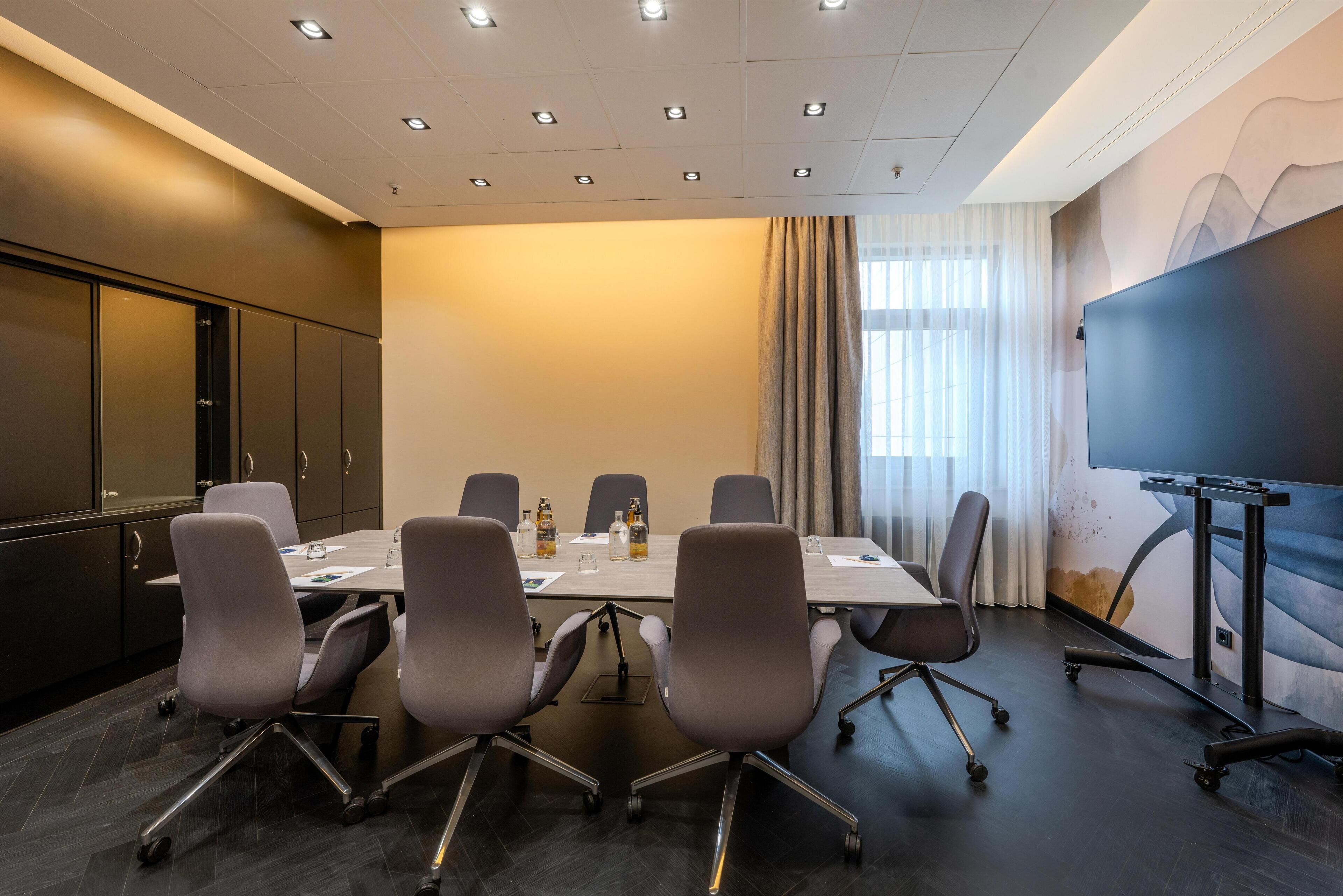 Une salle de conférence équipée d’une longue table, de six chaises, d’un écran de projection et d’une grande fenêtre avec des rideaux.
