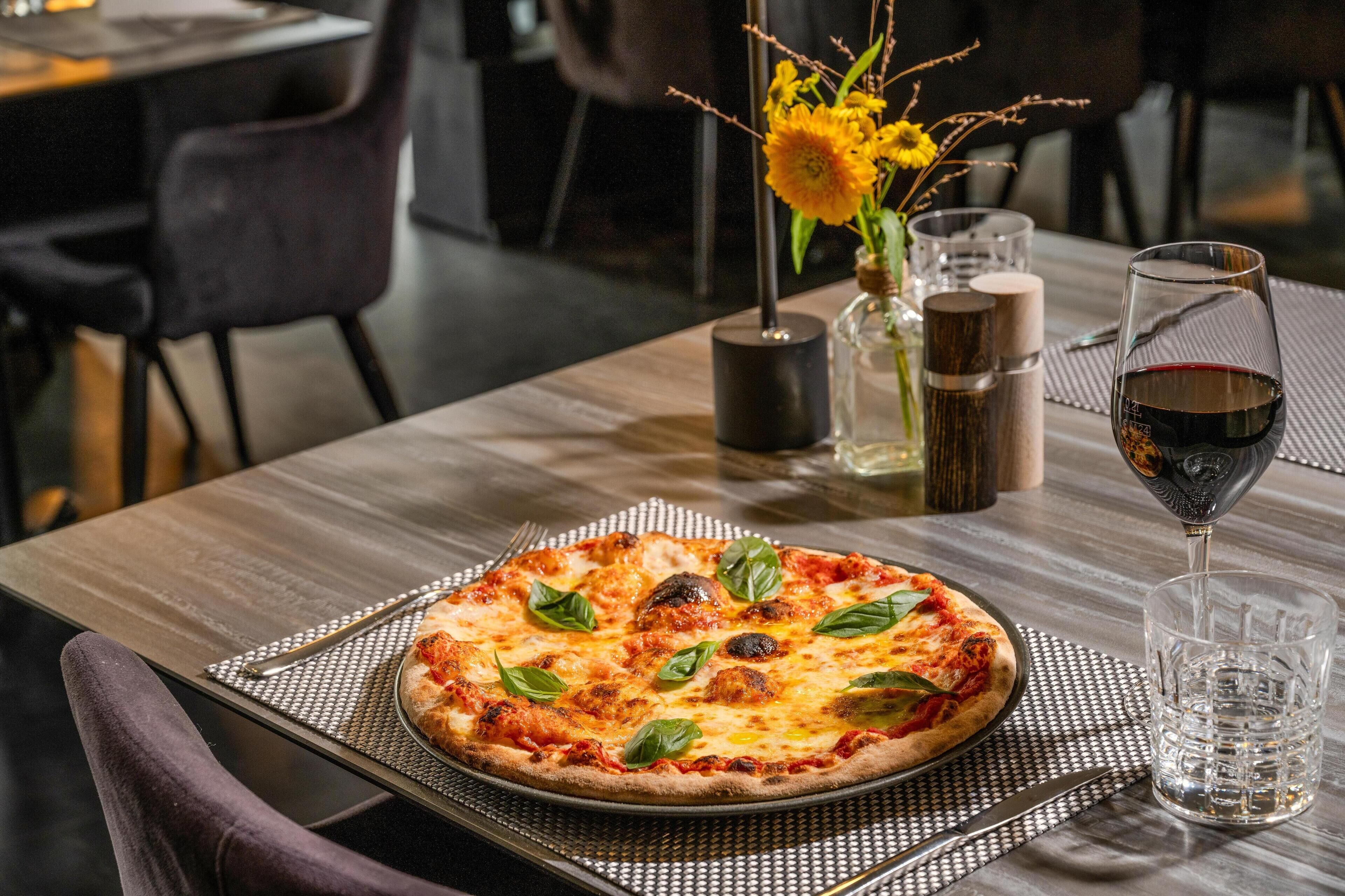 Eine Pizza auf einem Tisch, dazu ein Glas Rotwein und eine Vase mit Blumen.
